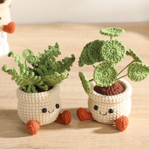 May include: Two crocheted plant pots with smiling faces, cream-coloured bodies, and small orange feet. One pot contains a green fern-like plant with curled fronds, while the other holds a green plant with round, flat, circular leaves.