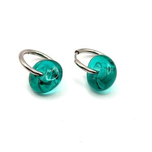 Puede incluir: Un par de pendientes de aro con aros de metal plateado pulido, cada uno adornado con una cuenta de vidrio turquesa redondeada y translúcida. Las cuentas tienen un acabado liso y brillante y cuelgan libremente de los aros metálicos.