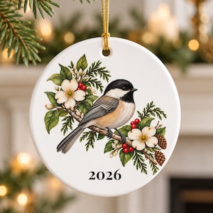 May include: A round white ceramic ornament featuring a black-capped chickadee perched on a branch with white dogwood flowers, green leaves, red berries, and pinecones. The year 2026 is printed in black at the bottom, and it hangs by a gold cord.