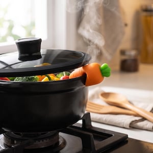 Può includere: Una pentola nera con coperchio in vetro e una clip in silicone a forma di carota per lo sfiato del vapore fissata sul bordo. La pentola contiene verdure colorate e il vapore fuoriesce dall'apertura, creando una scia visibile sopra.
