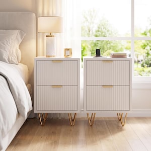 May include: Two white nightstands with two drawers each, featuring vertical ribbed textures, gold-toned horizontal drawer pulls, and gold-toned hairpin legs. The nightstands are rectangular in shape and stand on a light-colored wooden floor.