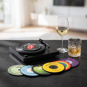 Puede incluir: Un tocadiscos negro con un disco de Rolling Tones en el plato, acompañado de una copa de vino y un vaso de whisky. Seis posavasos circulares diseñados como discos de vinilo con varios colores y etiquetas están alineados al frente.