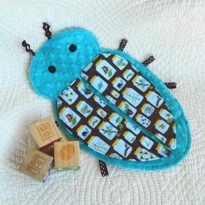 May include: A turquoise plush bug-shaped baby toy with black antennae and black dot accents. The bug's wings are patterned with jars containing insects and plants. Wooden blocks with etched designs are next to the toy.