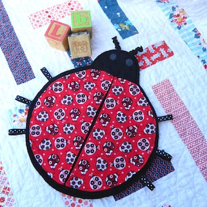 Op de afbeelding: Een rood en zwart lieveheersbeestje-vormig babyspeelgoed met zwarte lintjes. Het speelgoed is gemaakt van stof en heeft een zwart-wit stippenpatroon.