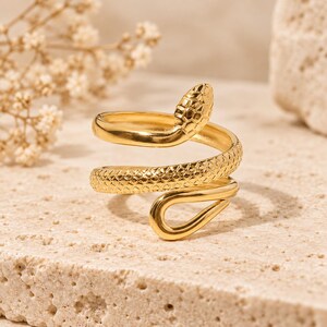 May include: A gold-toned ring shaped like a coiled snake. The ring features a textured, scale-like pattern along the body of the snake, with a smooth, rounded head and a tapered tail end that curves to form the open-ended band design.