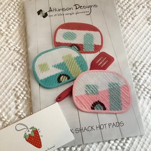 May include: Three colorful hot pads shaped like campers. The hot pads are pink, blue, and red with white accents. The hot pads are on a white wooden surface. The text "K SHACK HOT PADS" is printed on the surface.