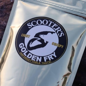 May include: A shiny gold metallic pouch featuring a circular black and white label. The label displays the text SCOOTER'S GOLDEN FRY, LIGHT, CRISPY, and UNFORGETTABLE TASTE! with a stylized black graphic of a fish shape integrated into the letter S.