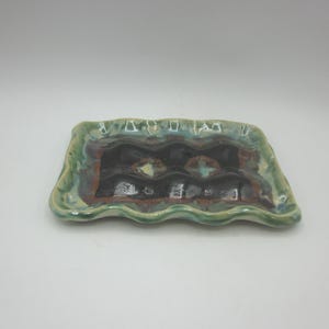 Peut inclure: Un plateau rectangulaire en céramique avec un bord ondulé. Le plateau a un bord vert et bleu, avec un centre marron foncé et noir. Le centre présente un motif décoratif avec des touches de jaune et turquoise. Le plateau mesure environ 20 cm de long.