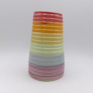 Könnte beinhalten: Eine zylindrische Keramikvase mit Regenbogenfarben. Die Vase ist vertikal mit Bändern aus Rot, Orange, Gelb, Grün, Blau und Lila gestreift. Die Vase hat eine strukturierte, gerippte Oberfläche.