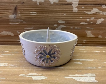 May include: A white ceramic ring holder with a blue and white floral design. The holder has a small, pointed spike in the center.