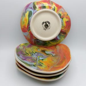 May include: A stack of three colorful ceramic plates with a marbled design in shades of orange, yellow, green, purple, and blue. The bottom plate has a white center with a black logo that says "Clay Kitten".
