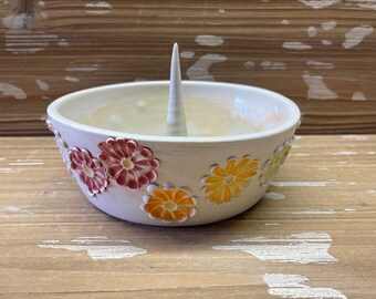 May include: White ceramic ring holder with a central spire and a decorative border of colorful flowers. The flowers are in shades of red, orange, and yellow.