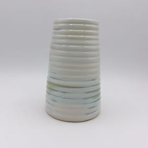 Könnte beinhalten: Weiße Keramikvase mit blauen und grünen Streifen. Die Vase hat eine zylindrische Form mit einem leicht ausgestellten oberen Rand.