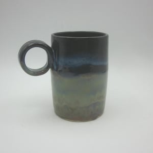 Könnte beinhalten: Eine hohe Keramik-Tasse mit einem einzigartigen kreisförmigen Griff. Die Tasse hat eine Farbverlaufs-Farbgebung, die von Dunkelblau oben nach Grün und Braun unten übergeht. Die Tasse ist etwa 13 cm hoch.