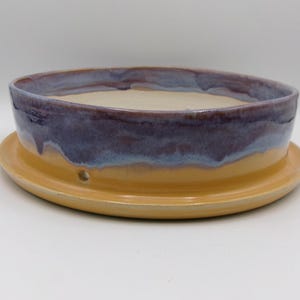 Op de afbeelding: Een gele keramische plantenpot met een paarse en blauwe glazuur. De pot heeft een schotel met drainagegaten.