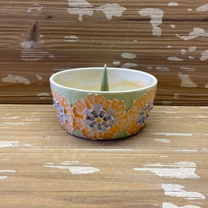 May include: A ceramic incense holder with a green and white floral design. The holder has a small cone-shaped opening in the center for incense.
