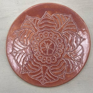Könnte beinhalten: Ein runder, terracottafarbener Keramikteller mit einem weißen, filigranen Blumendesign. Das Design zeigt eine zentrale Blume mit mehreren Blütenblättern und Blättern.