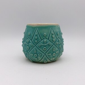 Può includere: Un vaso in ceramica color turchese con un motivo geometrico. Il vaso ha una forma arrotondata e una finitura liscia e smaltata.