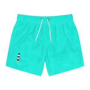 May include: Bright turquoise swim trunks featuring an elastic waistband with white drawstring cords. A small graphic of a blue and white striped surfboard standing upright on a patch of tan sand is positioned on the lower left leg area.