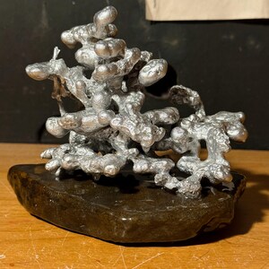 May include: A silver-colored metal sculpture with an irregular, organic, and porous structure resembling molten metal. The metallic form is mounted on a dark, smooth, flat, and irregularly shaped stone base, creating a contrast in texture and color.