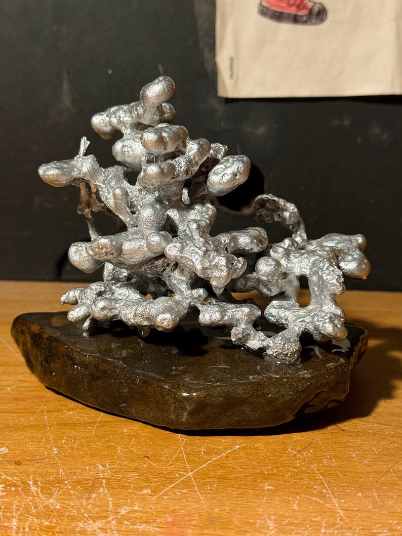 May include: A silver-colored metal sculpture with an irregular, organic, and porous structure resembling molten metal. The metallic form is mounted on a dark, smooth, flat, and irregularly shaped stone base, creating a contrast in texture and color.
