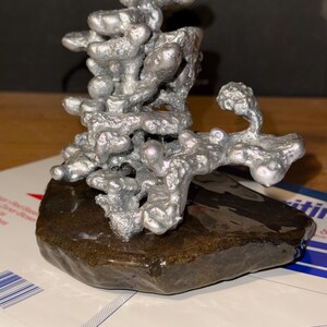 May include: A silver-colored, abstract metal sculpture with a porous, organic, and branching structure. The sculpture is mounted on a dark, smooth, flat, and irregularly shaped stone base, creating a contrast between the metallic and natural elements.