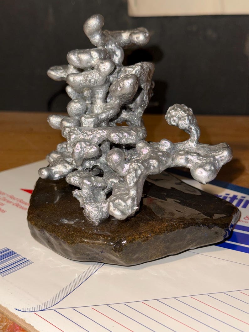 May include: A silver-colored, abstract metal sculpture with a porous, organic, and branching structure. The sculpture is mounted on a dark, smooth, flat, and irregularly shaped stone base, creating a contrast between the metallic and natural elements.