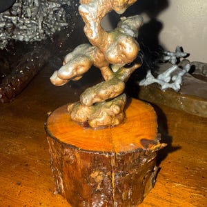 May include: A sculpture featuring a metallic, bronze-toned, organic, and twisted form mounted on a cylindrical, natural wood log base. The metallic structure has a textured, uneven surface with hints of silver and copper tones throughout the piece.
