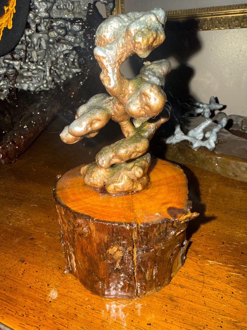 May include: A sculpture featuring a metallic, bronze-toned, organic, and twisted form mounted on a cylindrical, natural wood log base. The metallic structure has a textured, uneven surface with hints of silver and copper tones throughout the piece.