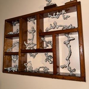 May include: A rectangular wooden wall shelf with multiple compartments containing irregular, silver-colored metal castings. The dark brown wooden frame features various sized cubbies, each displaying unique, organic-shaped metallic structures.