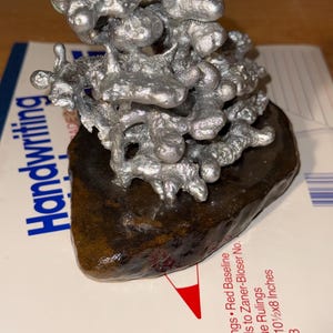 May include: A silver-colored, irregular, molten-style metal sculpture with a textured, organic shape. The sculpture is mounted on a dark brown, smooth, flat stone base. The object is positioned on a white paper surface with visible printed text.