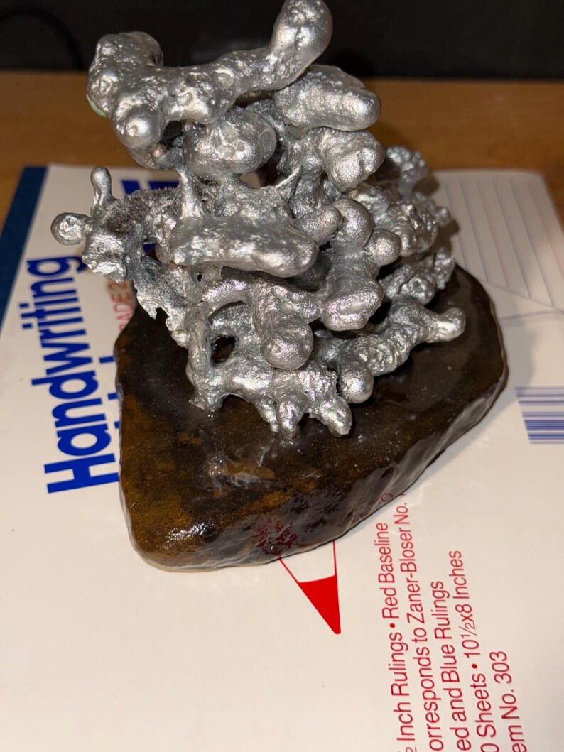 May include: A silver-colored, irregular, molten-style metal sculpture with a textured, organic shape. The sculpture is mounted on a dark brown, smooth, flat stone base. The object is positioned on a white paper surface with visible printed text.