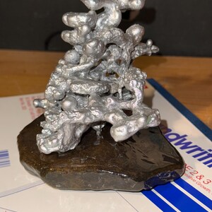 May include: A silver-colored, irregular, molten-looking metal sculpture with a porous, organic texture. The sculpture is mounted on a dark, smooth, flat, brown stone base, creating a contrast between the metallic top and the natural rock bottom.
