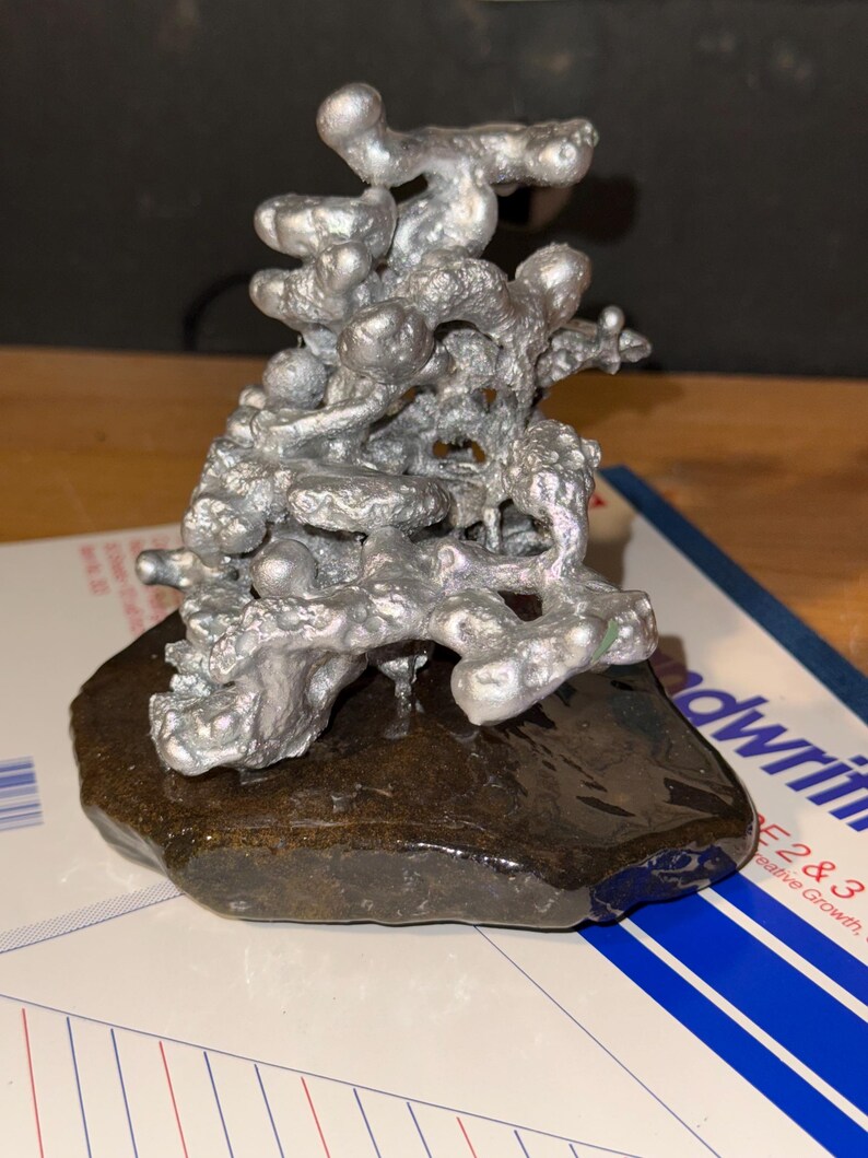 May include: A silver-colored, irregular, molten-looking metal sculpture with a porous, organic texture. The sculpture is mounted on a dark, smooth, flat, brown stone base, creating a contrast between the metallic top and the natural rock bottom.