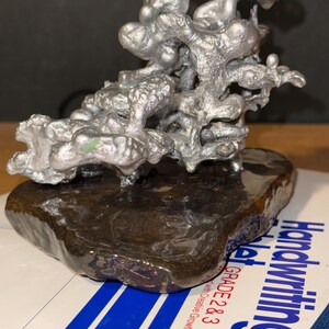 May include: A silver-colored, organic-shaped metal sculpture with a textured, branching structure mounted on a smooth, dark brown, polished stone base. The sculpture sits atop a white paper surface with blue lines and the text Handwriting Tablet Grade 2 & 3.