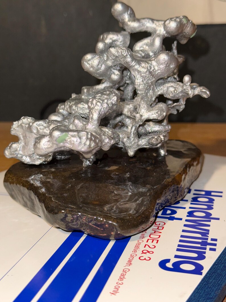 May include: A silver-colored, organic-shaped metal sculpture with a textured, branching structure mounted on a smooth, dark brown, polished stone base. The sculpture sits atop a white paper surface with blue lines and the text Handwriting Tablet Grade 2 & 3.
