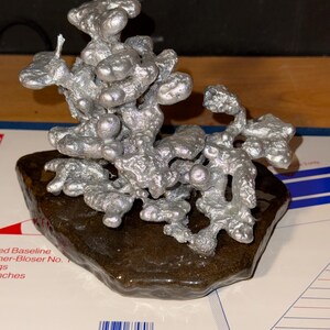 May include: A silver-colored, organic-shaped metal sculpture with a textured, bubbly surface. The sculpture is mounted on a dark brown, irregular, glossy base. The metal structure features multiple rounded, interconnected protrusions.