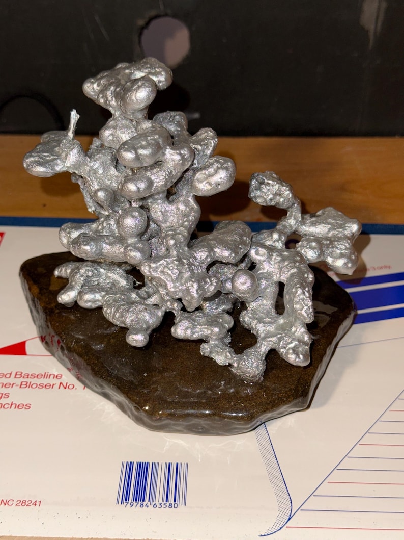 May include: A silver-colored, organic-shaped metal sculpture with a textured, bubbly surface. The sculpture is mounted on a dark brown, irregular, glossy base. The metal structure features multiple rounded, interconnected protrusions.