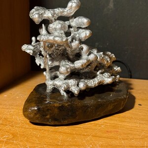 May include: A silver-colored metal sculpture with an organic, porous, and branching structure attached to a smooth, dark brown, flat stone base. The metallic portion features rounded, bulbous protrusions and irregular, interconnected shapes.