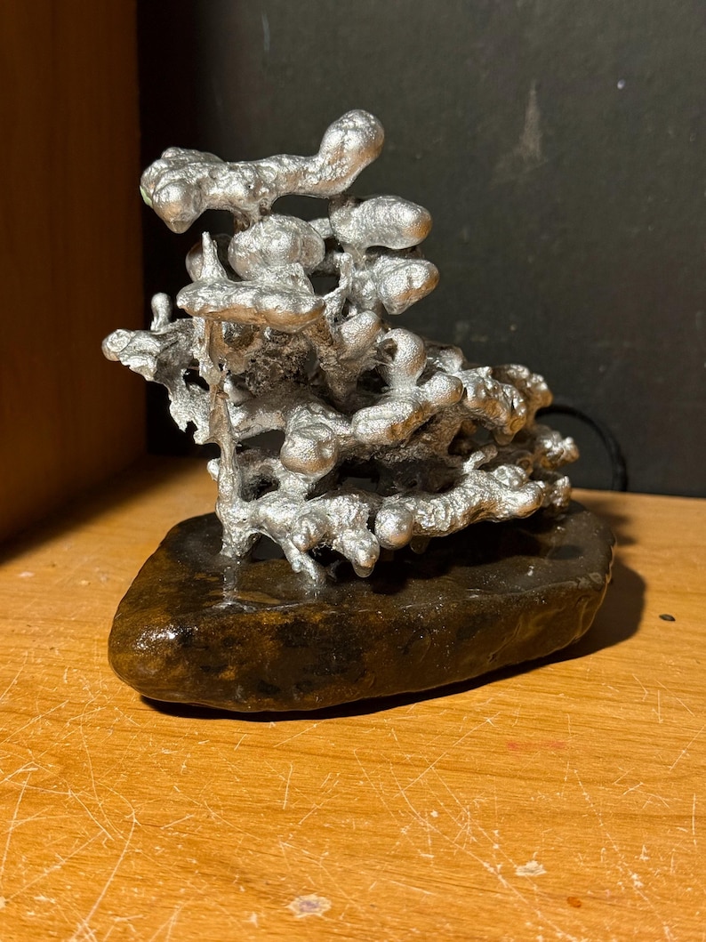 May include: A silver-colored metal sculpture with an organic, porous, and branching structure attached to a smooth, dark brown, flat stone base. The metallic portion features rounded, bulbous protrusions and irregular, interconnected shapes.