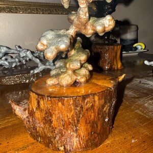 May include: A sculpture featuring a metallic, copper-toned, organic, and bulbous form mounted on a cylindrical, polished wooden log base. The upper structure has a textured, uneven surface with hints of green oxidation, contrasting with the smooth, warm brown wood grain of the base.