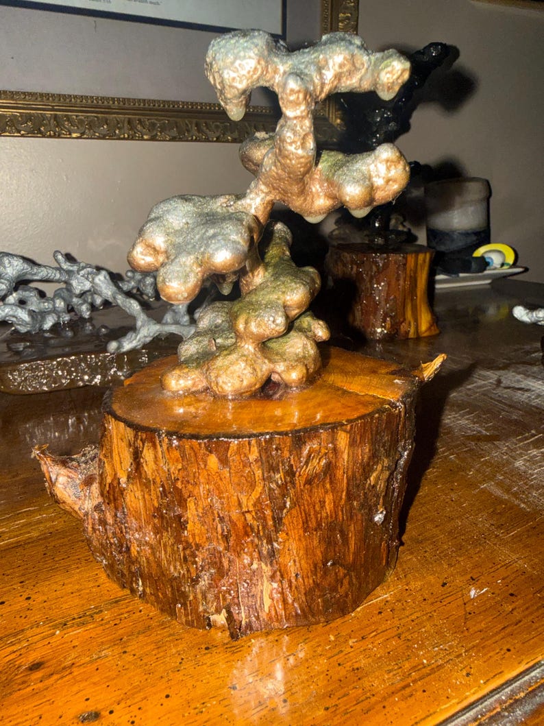 May include: A sculpture featuring a metallic, copper-toned, organic, and bulbous form mounted on a cylindrical, polished wooden log base. The upper structure has a textured, uneven surface with hints of green oxidation, contrasting with the smooth, warm brown wood grain of the base.