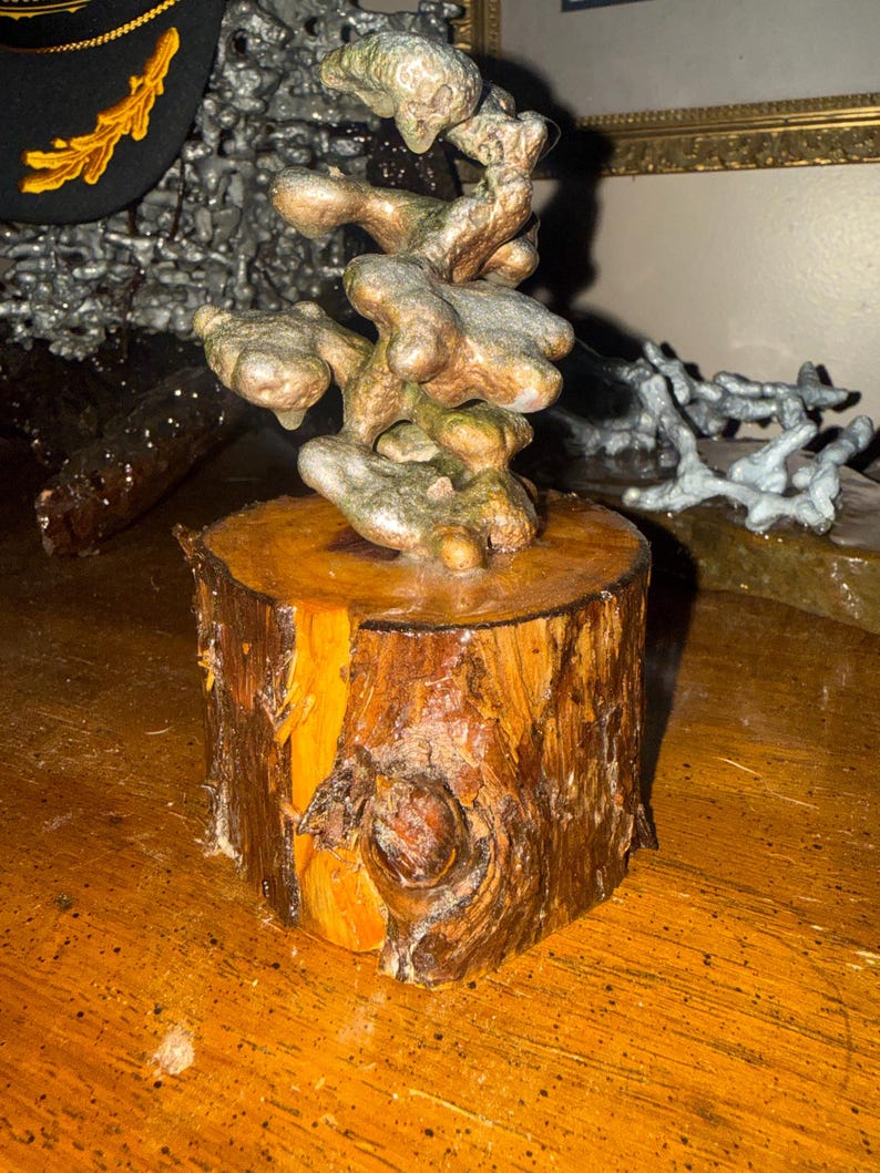 May include: A sculpture featuring a metallic, organic, and textured form with bronze and silver tones mounted on a cylindrical wooden base. The base displays natural wood grain, bark edges, and a prominent knot, providing a sturdy foundation for the abstract, multi-lobed metal structure above.