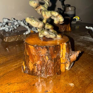 May include: A decorative sculpture featuring a textured, organic, greenish-brown form mounted atop a cylindrical wooden base with visible bark and a polished, amber-toned finish. The piece displays a gnarled, branching structure with rounded, bulbous segments that rise vertically from the center of the wood block.