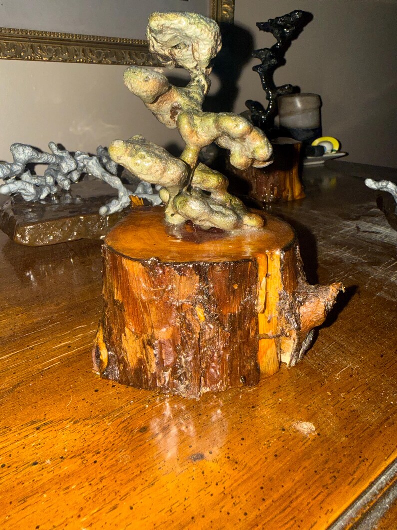 May include: A decorative sculpture featuring a textured, organic, greenish-brown form mounted atop a cylindrical wooden base with visible bark and a polished, amber-toned finish. The piece displays a gnarled, branching structure with rounded, bulbous segments that rise vertically from the center of the wood block.