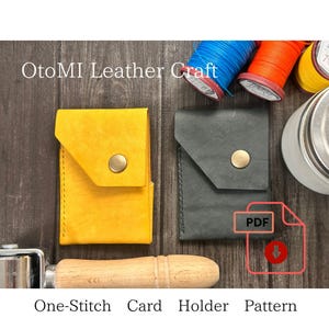 May include: Two leather card holders, one yellow and one dark gray, each featuring a single brass snap button closure. The text OtoMI Leather Craft and One-Stitch Card Holder Pattern is visible, along with spools of thread and a wooden tool.