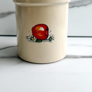 May include: A cylindrical beige ceramic crock featuring a hand-painted illustration of a bright red apple centered on the front, flanked by two small white daisies with green leaves. The vessel has a smooth finish and a thick, rounded rim.