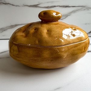 May include: A glossy, golden-brown ceramic lidded container shaped like a potato. The surface features an irregular, textured finish with small indentations and a smooth, reflective glaze. A rounded knob sits atop the lid, mimicking the appearance of a natural potato skin with its earthy, warm tones and organic, uneven form.