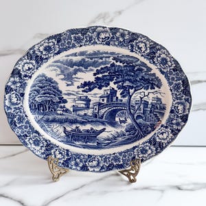 May include: An oval ceramic platter featuring a blue and white transferware pattern. The center depicts a landscape with a stone bridge, castle ruins, and a boat on a river. The scalloped rim is decorated with a dense floral border.