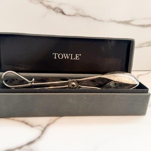 May include: A silver-toned metal serving tong with a spoon-shaped end and a looped handle, featuring a central circular hinge detail. The utensil is presented inside a rectangular dark gray storage box with the brand name TOWLE printed in white.